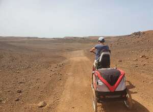 Frau mit Fahrrad, Kindersitz und Anhänger fährt durch eine wüstenartige Landschaft